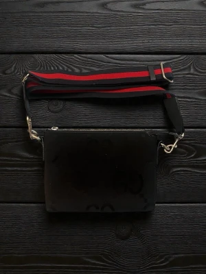 Svart axelväska från Gucci - Snygg svart axelväska från Gucci med diskret GG-mönster i tyget. Väskan har en bred axelrem i svart och rött med silverfärgade metalldetaljer. Perfekt för dig som vill ha en stilren och trendig accessoar.