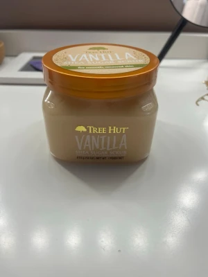 Tree Hut Vanilla Shea Sugar Scrub - Vaniljdoftande shea sugar scrub från Tree Hut i en burk med orange lock. Innehåller 510 g och har en krämig, ljusbeige färg. Perfekt för exfoliering och mjukgörande av huden med en söt vaniljdoft. Bara använd tre gånger 