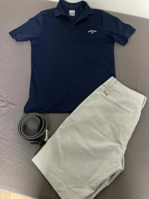 Ljusgråa shorts från Puma med bälte - Säljer en snygg pikétröja från Callaway i mörkblått. Passar tillsammans med ett par stilrena ljusgråa shorts från Puma med klassisk passform och Puma-logga ovanför bakfickan i storlek 28. Shortsen har en diskret design och kommer med ett grått flätat bälte från Jack & Jones (längd 90). Perfekta för sportiga aktiviteter och varma dagar. 