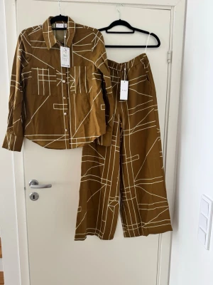 Brunt set med grafiskt mönster från Vila - Snyggt tvådelat set från Vila i brunt med vitt grafiskt linjemönster. Skjortan har krage, knappar framtill och bröstficka. Byxorna är vida med resår i midjan för skön passform. Perfekt för dig som gillar trendiga och bekväma plagg med unik design. Nypris 1 000kr