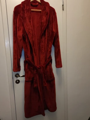 Mjuk vinröd morgon/badrock i fleece - Supermjuk vinröd morgonrock i fleece med långa ärmar och knytskärp i midjan. Perfekt för att chilla hemma och hålla sig varm. Klassisk modell med sjalkrage och två fickor framtill. Snygg och bekväm att dra på sig direkt på morgonen eller efter duschen. L/XL