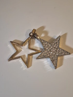 Halsband med dubbla stjärnhängen - Snyggt halsband med två stjärnformade hängen i metall, varav en är öppen och den andra är täckt med glittrande stenar. Perfekt för dig som gillar unika och trendiga smycken med lite extra bling. Passar till många olika stilar och outfits.
