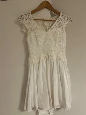 Vit spetsklänning med v-ringning - Superfin vit kortklänning med spets upptill och v-ringning både fram och bak. Klänningen har ärmlös design med spetsdetaljer på axlarna och en flowy kjol i lätt material, använd 1 gång och har inga defekter💕 ( pris kan diskuteras)