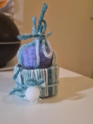 Blåvit stickad minihatt med boll - En liten handgjord minihatt i blått, vitt och lila garn med dekorativ boll och snören på toppen. Hatten har ett randigt mönster och är tillverkad i mjukt ullgarn. Perfekt som accessoar eller dekoration för dig som gillar unika detaljer.