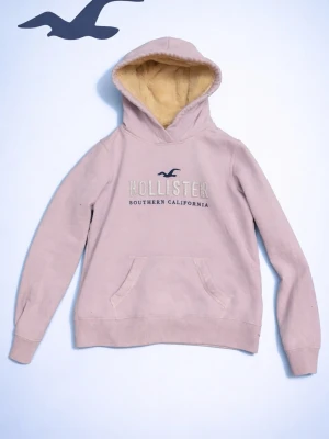 Ljuslila fur hoodie från Hollister - Mysig ljuslila hoodie från Hollister med broderad logga och texten 'Southern California' på bröstet. Hoodien har en stor ficka framtill och en fodrad huva i beige teddy-material. Perfekt för chill dagar och streetwear-stil.