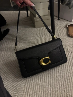 Svart axelväska  - Svart axelväska i strukturerat material med guldfärgad spänndetalj framtill. Slank och stilren modell med lockstängning och justerbar axelrem. Insida med flera fack som gör det enkelt att hålla ordning. Funkar lika bra till vardag som kväll.