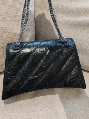 Svart quiltad handväska Balenciaga - Snygg svart quiltad handväska från Balenciaga med kedjeremmar och ett stort B-spänne framtill. Väskan har en unik form och är tillverkad i glansigt material. 31x20x9cm