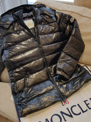 Svart dunjacka från Moncler - Snygg svart dunjacka från Moncler med glansig finish och klassisk Moncler-logga på ärmen. Jackan har dragkedja framtill, hög krage och quiltad design. Riktigt fint tillverkad!. Jag fick den som present, sitter fint, jag är 175cm, 80kg. 