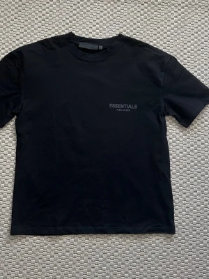 Essentials Fear of God t-shirt - Säljer en svart t-shirt från Fear of God Essentials med tryck fram och stort logotyp-tryck på ryggen. Klassisk passform med rund hals och korta ärmar. Perfekt för dig som gillar streetwear och enkel stil.