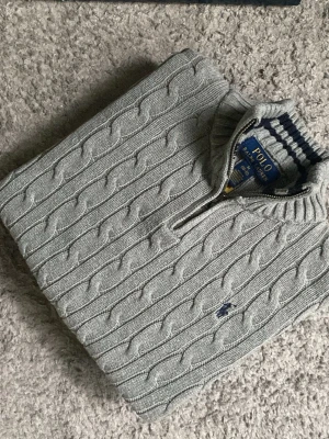 Grå kabelstickad tröja Polo Ralph Lauren - Kabelstickad Grå  tröja från Polo Ralph Lauren med halv dragkedja och hög krage. Har också den klassiska broderade loggan på bröstet. Perfekt för en stilren och avslappnad look.