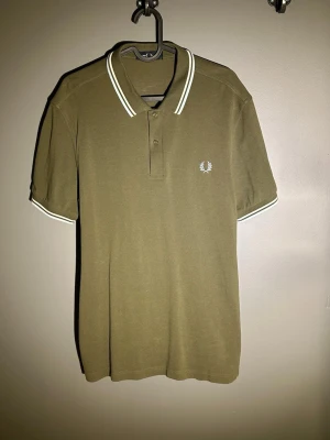 Olivgrön pikétröja från Fred Perry - Klassisk olivgrön pikétröja från Fred Perry med vit broderad logga på bröstet. Tröjan har vitrandiga detaljer på krage och ärmslut samt knappar framtill. Tillverkad i mjuk bomull och har en normal passform med korta ärmar.