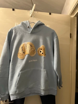 Blå hoodie från Palm Angels med nalle - Säljer en ljusblå hoodie från Palm Angels med en unik applikation av en nallebjörn på bröstet. Hoodien har huva, magficka och är gjord i mjukt material. Perfekt för dig som gillar streetwear och vill sticka ut med en cool detalj framtill.