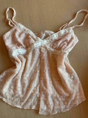 Beige spetslinnetopp med tunna band - Söt ljus ros linnetopp med tunna axelband och fina spetsdetaljer längs bysten och under bysten. Toppen har ett lätt transparent tyg med broderade blommor och en luftig passform. Perfekt för varma dagar eller lager på lager.