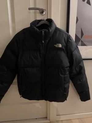 Svart pufferjacka från The North Face - Säljer en svart pufferjacka från The North Face med hög krage och dragkedja framtill. Jackan har logga på bröstet och ryggen, samt breda, vadderade paneler som ger en riktigt fet siluett. Perfekt för kalla vinterdagar och riktigt snygg streetstil. Även såklart äkta köpt på Stadium för ungefär 2 år sedan sparsamt använd och jackan passar för både könen. Nypris- 2700 