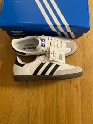 Adidas Samba vita och svarta sneakers - Klassiska Adidas Samba sneakers i vitt läder med svarta ränder och brun gummisula. Skon har detaljer i grå mocka på tån och guldfärgad SAMBA-text på sidan. Perfekt för dig som gillar retrostil och streetwear.