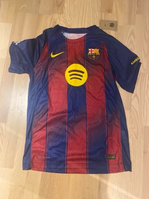 FC Barcelona matchtröja Nike - Säljer en FC Barcelona matchtröja från Nike med klassiska blå och röda vertikala ränder. Tröjan har klubbmärke, Nike-logga och Spotify som sponsor framtill. Champions League-märke på ärmen och gul detalj på andra ärmen. Perfekt för fotbollsfans!