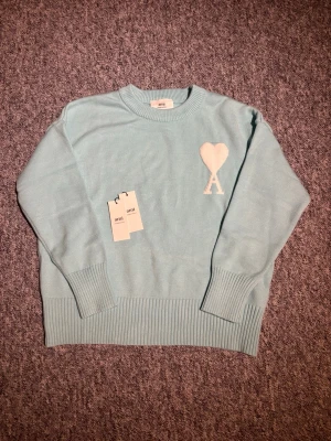 Ljusblå stickad tröja från Ami Paris S - Ljusblå stickad tröja från Ami Paris med rund halsringning och ribbade muddar. På bröstet finns ett vitt hjärta med bokstaven A, typiskt för märket. Tröjan har en avslappnad passform och är perfekt för lager-på-lager-stil. Pris kan diskuteras 😊