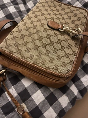 Axelväska från Gucci (äkta) - Vintage och snygg beige axelväska från Gucci med klassiskt GG-mönster i canvas och detaljer i äkta brunt läder. Väskan har justerbar axelrem, dragkedja och dekorativ flätad läderdetalj. Perfekt för dig som gillar lyxiga accessoarer med ikonisk design. Det finns också äkthetsbevis och väskan har blivit verifierad av Vestaire Collective. 