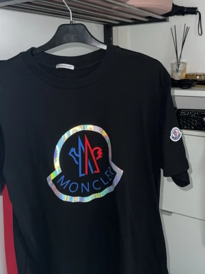 Svart Moncler t-shirt med holografiskt tryck - Säljer en svart t-shirt från Moncler med ett stort holografiskt logotryck på bröstet och Moncler-märke på ärmen. T-shirten har rund hals och korta ärmar. Perfekt för dig som vill sticka ut med en exklusiv streetwear-look.