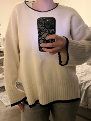 Vit oversized stickad tröja - Säljer en vit oversized stickad tröja med svarta kontrastkanter vid hals, ärmslut och nederkant. Tröjan har bred ribbstickning nedtill och på ärmarna samt en avslappnad passform. Perfekt för dig som gillar en enkel men snygg look.