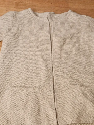 Beige strukturerad cardiganjacka - Stilren beige cardiganjacka med rund halsringning och öppen front. Jackan har två diskreta fickor framtill och är tillverkad i ett mjukt, strukturerat material med stickad känsla. Perfekt för lager på lager och enkel att matcha med olika outfits.