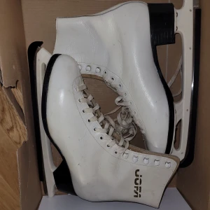 Vita konståkningsskridskor Jofa - Säljer ett par klassiska vita konståkningsskridskor från Jofa. Skridskorna har högt skaft, snörning framtill och svart sula. Bladen är i metall och skon är tillverkad i syntetmaterial med Jofa-logga på sidan. Perfekta för isbanan eller träning.