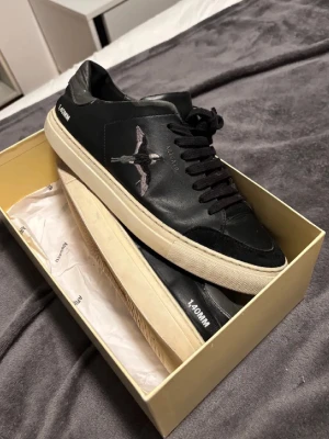 Svarta Axel Arigato sneakers med fågel - Snygga svarta sneakers från Axel Arigato i läder med broderad fågel på sidan och vit sula. Klassisk låg modell med svarta skosnören och diskret logga på sidan. Perfekta för dig som gillar stilrena och trendiga skor. Allt på bilderna ingår ( Arigato påse, box & skorna)