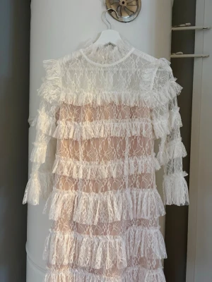 By Malina klänning - Superfin By Malina vit spetsklänning med volanger. Klänningen har långa ärmar och hög hals, samt dragkedja i ryggen. Perfekt för dig som vill sticka ut med en romantisk och trendig look. Passar perfekt till studenten/konfirmationen!🤍