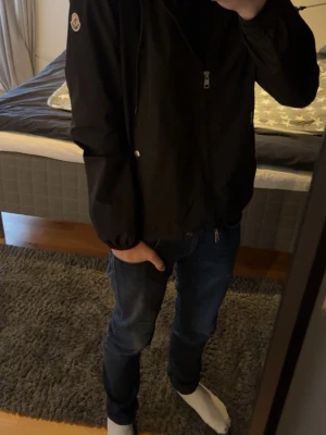 Svart vindjacka från Moncler❤️🖤 - Snygg svart vindjacka från Moncler med dragkedja framtill, huva med röd och vit rand samt två fickor med tryckknappar. Jackan har elastiska muddar vid ärmsluten och en klassisk Moncler-logga på ärmen. Perfekt för blåsiga dagar och enkel att matcha. Perfekt till kommande Vår👍🌟