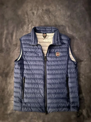 Väst från Colmar - Snygg blå dunväst från Colmar med quiltad design och hög krage. Västen har två dragkedjeförsedda fickor framtill och en liten Colmar-logga på bröstet. Perfekt att slänga över en hoodie eller tröja för extra värme. Materialet är lätt och smidigt.