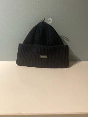 Mörkblå mössa från Sätila - Snygg blå ribbstickad mössa från Sätila med uppvikt kant och en liten metallbricka framtill. Perfekt accessoar för kalla dagar och enkel att matcha med olika outfits.