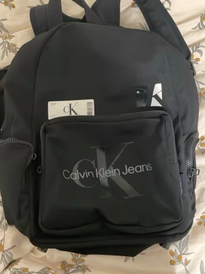Svart ryggsäck Calvin Klein Jeans - Snygg svart ryggsäck från Calvin Klein Jeans med stort cK-logotyptryck på frontfickan. Ryggsäcken har vadderade axelband, flera fack med dragkedja och sidofickor i mesh. Perfekt för skolan eller stan och rymmer allt du behöver.