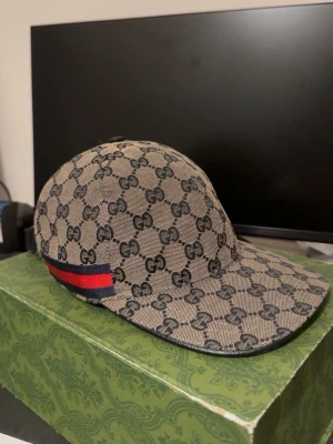 Gucci GG Supreme keps med röd-blå rand - Snygg keps från Gucci i beige och marinblått GG Supreme-mönster. Kepsen har en klassisk böjd skärm och en ikonisk röd-blå rand på sidan. Tillverkad i canvas med justerbart band bak och mörkblått innerfoder. Perfekt för dig som gillar exklusiva accessoarer.