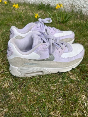 Nike Air Max lila och vita sneakers - Nike Air Max sneakers i lila och vitt med grå detaljer. Skorna har en klassisk låg siluett, mesh och mocka-paneler samt den ikoniska Air Max-sulan för extra komfort. Swoosh-logga på sidan och snörning framtill.