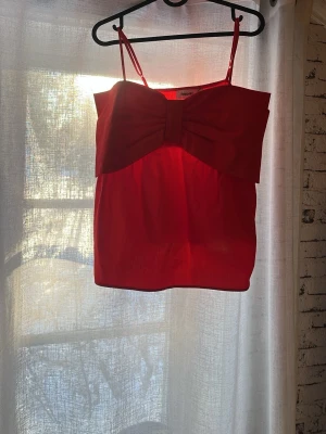 Röd topp - Supersnygg röd topp från NA-KD med tunna axelband och en stor rosett framtill som statement-detalj. Toppen har en croppad passform och är tillverkad i ett lätt, vävt material som känns bekvämt mot huden. Perfekt för dig som vill sticka ut! Helt ny med prislapp kvar 