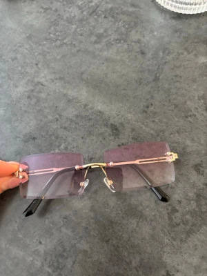 Rosa rektangulära solglasögon - Säljer ett par trendiga solglasögon med rektangulära, ljusrosa glas och tunna guldfärgade bågar. Glasen är utan ram och har en modern, stilren look. Perfekta för dig som vill sticka ut med en cool accessoar.