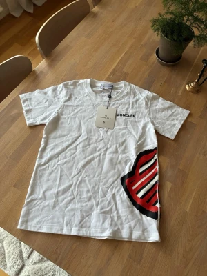 Vit Moncler t-shirt med logga - Snygg vit t-shirt från Moncler med svart tryckt logga på bröstet och en stor röd, svart och vit Moncler logga på sidan. Klassisk rund halsringning och korta ärmar. Tillverkad i mjuk bomull för en skön känsla.