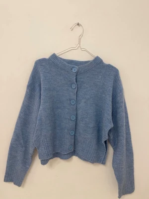 Ljusblå cropped kofta med knappar - Mysig ljusblå stickad kofta med stora matchande knappar framtill och ribbad nederkant. Har även coola slitsar på sidorna, som ger extra stilpoäng. Rak passform. Är strl M men passar mig som brukar ha XS/S