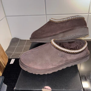 Bruna UGG Tasman II tofflor - Mysiga bruna UGG Tasman II tofflor i mocka med mjuk fårskinnsinsida och dekorativt mönster runt öppningen. Klassisk slip-on modell med rejäl sula som funkar både inne och ute. Perfekta för chill dagar hemma eller när du vill ha det extra bekvämt.