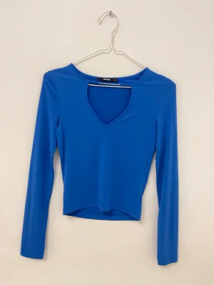 Blå långärmad topp med v-ringning - Snygg blå långärmad topp med v-ringad hals och croppad passform. Har en glitter detalj vid halsen. Perfekt att styla med jeans eller kjol för en trendig look. Tunt och stretchigt material som sitter skönt på kroppen.