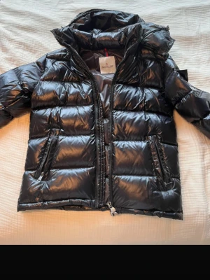 Svart dunjacka från Moncler - Snygg svart dunjacka från Moncler med glansig finish och klassisk puffer-design. Jackan har två dragkedjeförsedda fickor, hög krage och avtagbar huva. 