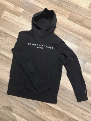 Svart hoodie från Tommy Hilfiger - Snygg svart hoodie från Tommy Hilfiger med klassisk logga och text på bröstet. Hoodien har en stor huva och magficka framtill. Inga prisförslag är dumma!
