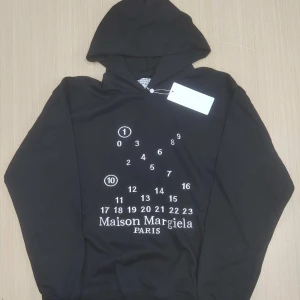 Maison Margiela svart hoodie med tryck - Svart hoodie från Maison Margiela med ikoniskt vitt siffertryck och logga på bröstet. Klassisk huva, långa ärmar och ribbade muddar. Mjuk bomullskänsla och minimalistisk design, perfekt för dig som gillar streetwear med lyxig twist.