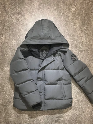  Canada Goose macmillan  - Snygg mörkgrå pufferjacka från Canada Goose med stor huva och patch-logga på ärmen. Jackan har dragkedja och tryckknappar framtill samt två sidofickor. Perfekt för kalla vinterdagar och riktigt varm. Pris går att diskutera så det är bara att höra av sig för de lilla minsta😊