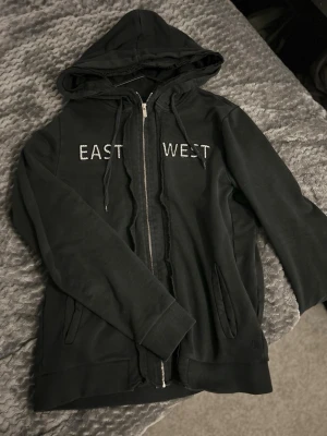 Svart hoodie från Les Benjamins - Svart hoodie med dragkedja från Les Benjamins. På bröstet står det 'EAST WEST' i vita bokstäver. Hoodien har två fickor framtill, justerbar huva med snören och ribbade muddar. Perfekt för en chill och avslappnad stil.