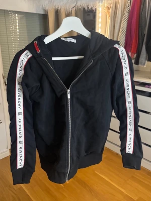  Givenchy Hoodie - Svart hoodie från Givenchy med dragkedja framtill och huva. Snygga vita band med Givenchy-logga längs båda ärmarna och röda detaljer vid axlarna. Tillverkad i mjukt bomullsmaterial och har ribbade muddar vid ärmslut och nederkant. Perfekt för en trendig streetwear-look.Dm för pris
