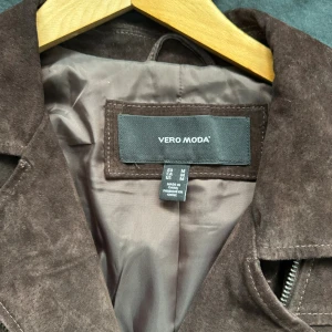 Brun skinnjacka från Vero Moda - Snygg brun skinnjacka från Vero Moda i bikerstil med flera silverfärgade dragkedjor och tryckknappar. Jackan har bred krage och markerade sömmar som ger en cool look. Perfekt för dig som vill ha en trendig och tidlös jacka.