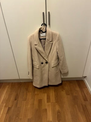 Beige kappa från Stradivarius - Stilren beige kappa från Stradivarius med två knappar framtill och klassisk krage. Kappan har två fickor och är fodrad för extra komfort. Perfekt för dig som vill ha en enkel men snygg look under höst och vinter.