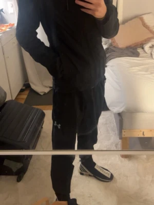 Svart träningsset från Under Armour - Säljer ett svart träningsset från Under Armour med matchande jacka och byxor. Setet har en sportig look med dragkedja på tröjan och diskret logga på byxan. Perfekt för gymmet eller löprundan. Byxorna - S, Jackan XS