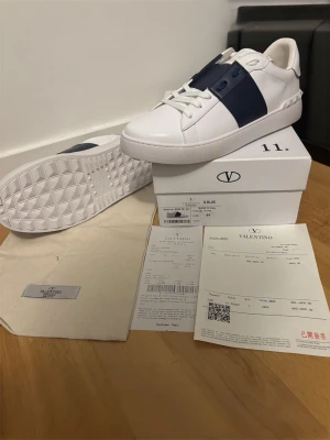 Valentino vita sneakers med blå detalj - Snygga vita sneakers från Valentino med bred mörkblå remsa över sidan. Skorna har klassisk låg profil, snörning och är tillverkade i slätt skinn. Perfekta för dig som vill ha en stilren och exklusiv look.